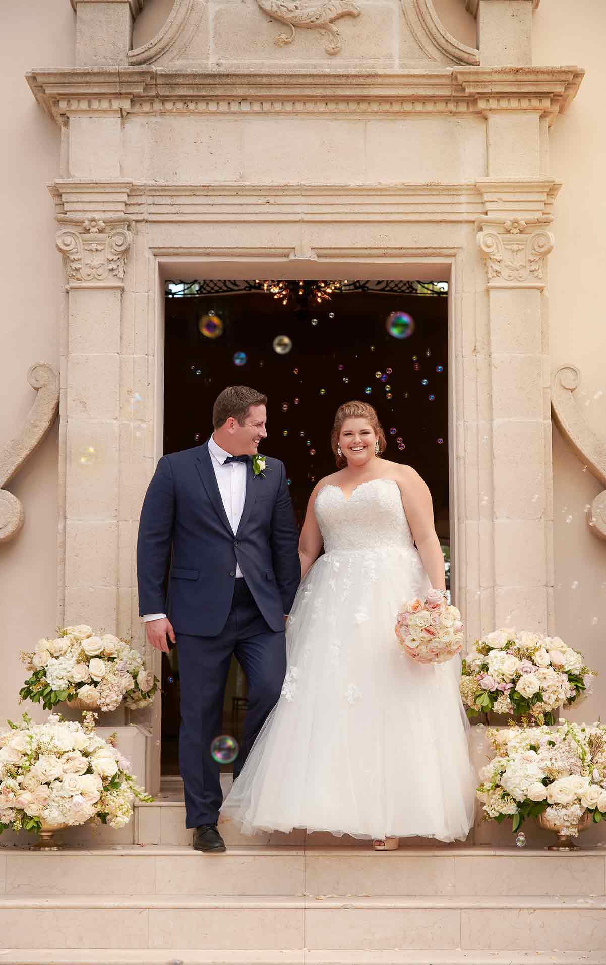 Eine Braut in einem atemberaubenden Hochzeitskleid steht mit ihrem Bräutigam auf Steinstufen, umgeben von weißen Blumen und Seifenblasen. Sie hält einen Blumenstrauß in der Hand, während er sie neben einem verschnörkelten Portal anlächelt. Beide strahlen an ihrem besonderen Tag vor Freude.