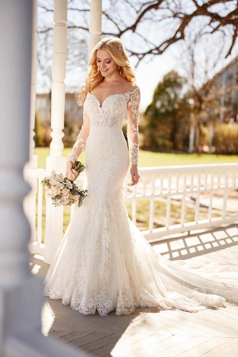 Eine Braut in einem schulterfreien Hochzeitskleid aus Spitze mit langen Ärmeln hält einen Blumenstrauß und lächelt, während sie auf einer sonnenbeschienenen Veranda mit Bäumen und Grünpflanzen im Hintergrund steht.
