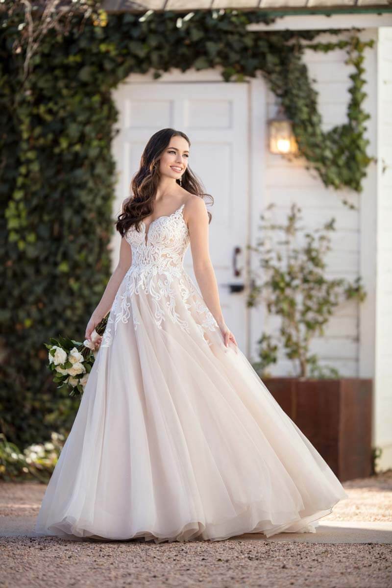 Eine lächelnde Braut in einem eleganten Hochzeitskleid, verziert mit Spitze, hält ihren Blumenstrauß und steht im Freien vor einem weißen Gebäude mit grüner Efeuwand.