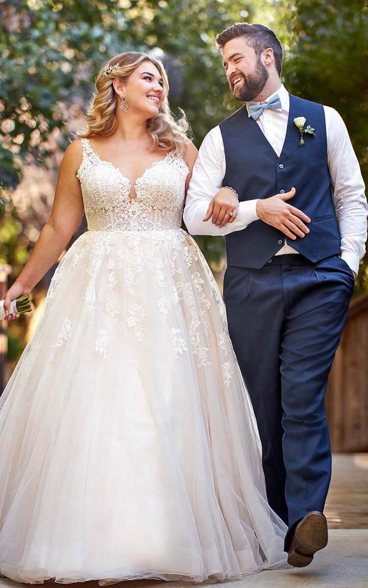 Eine Braut in einem weißen Brautkleid mit Spitzenbesatz und ein Bräutigam in einem marineblauen Anzug mit Weste gehen im Freien spazieren, lächeln sich freudig an und halten sich in den Armen, während im Hintergrund Grünpflanzen zu sehen sind.