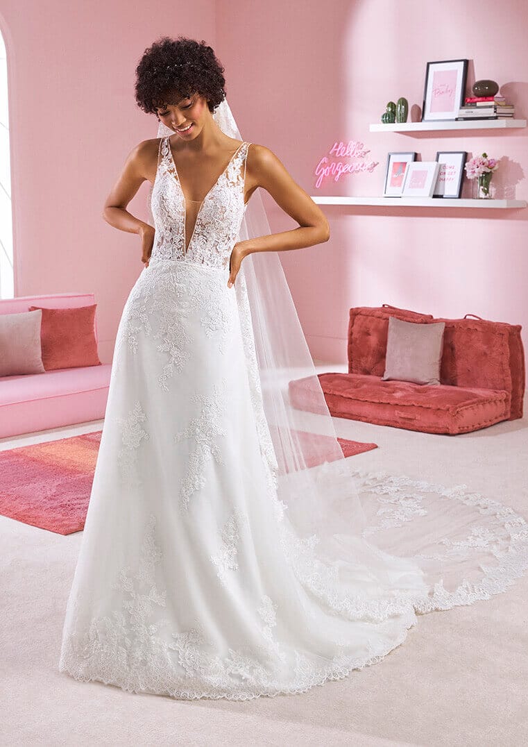 Eine lächelnde Frau, die ein weißes Spitzenbrautkleid mit langer Schleppe und durchsichtigem Mieder trägt, steht in einem rosafarbenen Raum mit moderner Einrichtung, darunter eine rosafarbene Couch, Kissen, gerahmte Kunst und ein Hello Gorgeous-Schild.