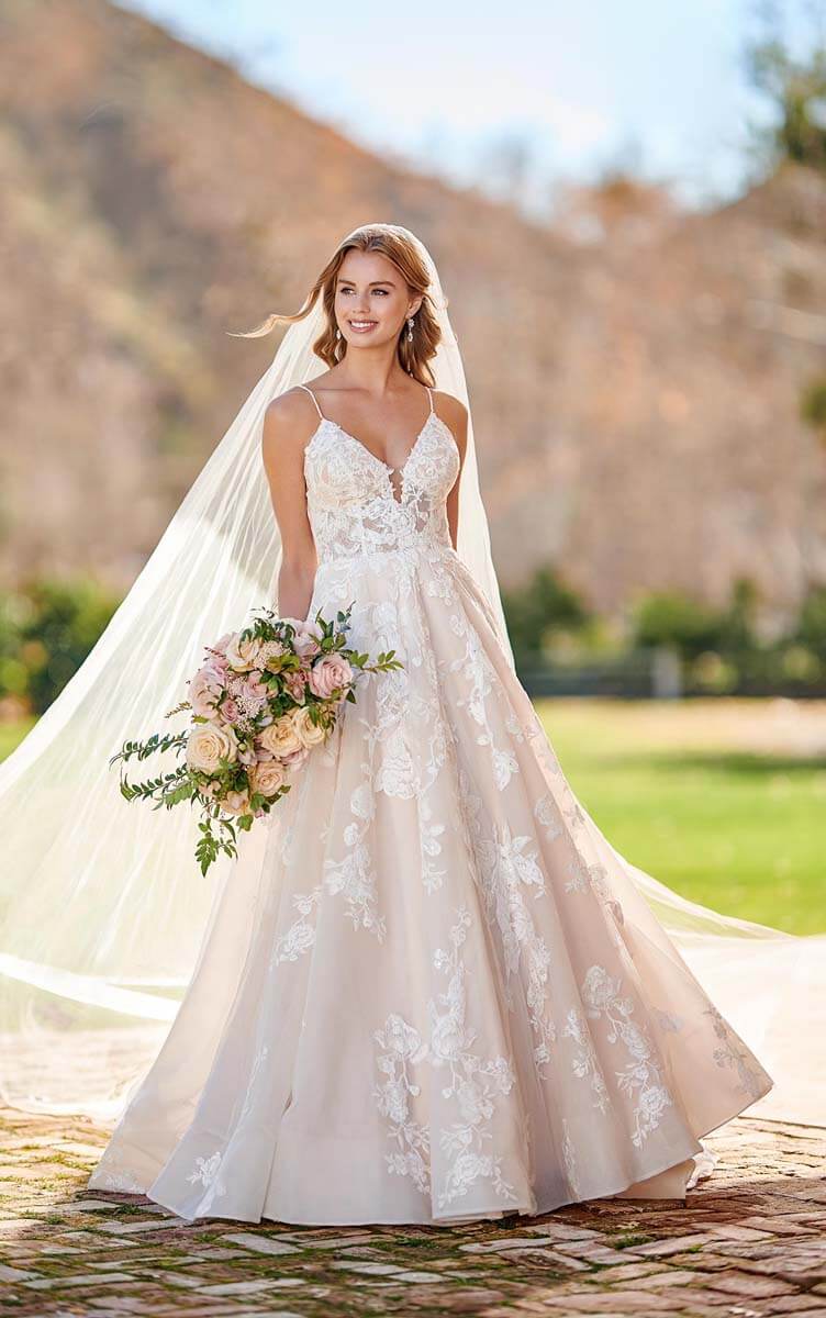 Eine lächelnde Braut in einem Brautkleid aus floraler Spitze und mit langem Schleier steht im Freien und hält einen Strauß aus rosa und weißen Blumen. Im Hintergrund sind verschwommene Bäume und Sonnenlicht zu sehen, die die Eleganz des modernen Brautmodens unterstreichen.