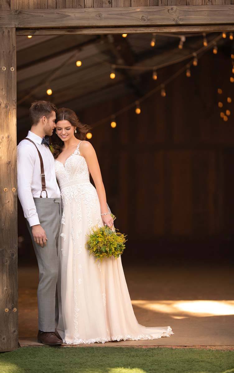 Eine Braut in einem zarten Hochzeitskleid mit einem gelben Blumenstrauß steht neben einem Bräutigam in grauen Hosen und Hosenträgern am Eingang einer Scheune, während im Hintergrund sanfte Lichterketten leuchten.