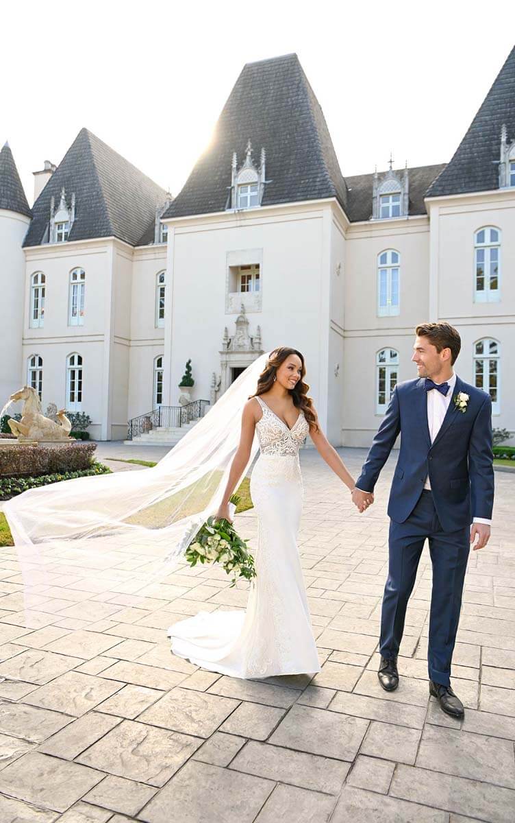 Eine Braut in einem atemberaubenden Brautkleid und Schleier hält die Hand des Bräutigams in einem blauen Anzug und lächelt einander an, während sie vor einem großen, eleganten Gebäude im Stil eines Schlosses spazieren gehen.