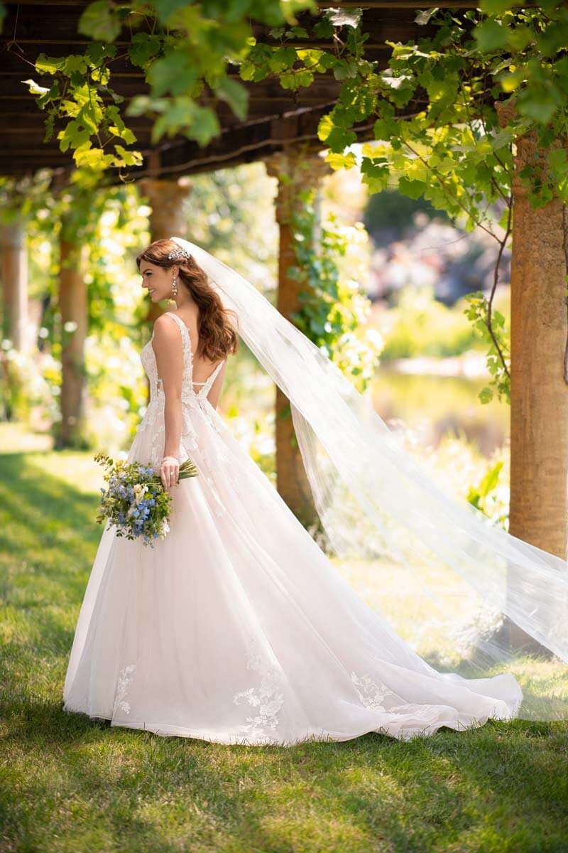 Eine Braut im Brautkleid mit langem Schleier steht im Freien unter einer begrünten Pergola, hält einen Blumenstrauß in der Hand und blickt an einem sonnigen Tag über ihre Schulter.