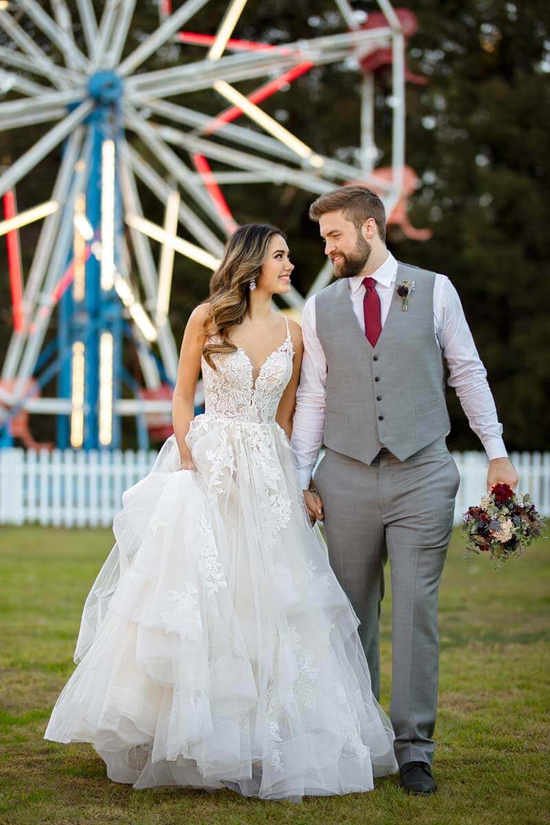 Eine Braut in einem weißen Brautkleid und ein Bräutigam in einem grauen Anzug mit roter Krawatte halten sich an den Händen und lächeln sich an, während sie auf einer Wiese mit einem Riesenrad im Hintergrund stehen.