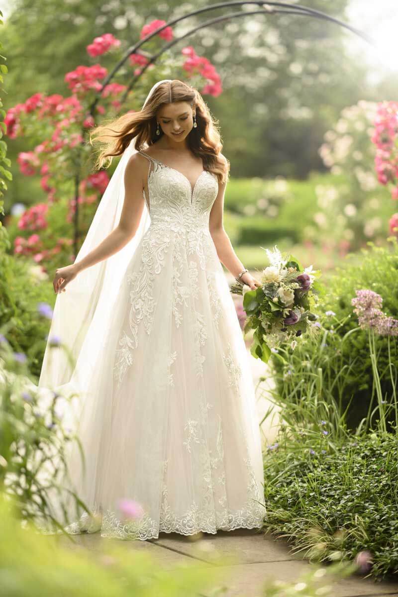 Eine lächelnde Braut in einem weißen Spitzen-Hochzeitskleid hält einen Blumenstrauß und steht auf einem Gartenweg, umgeben von üppigem Grün und blühenden rosa Blumen an einem sonnigen Tag.