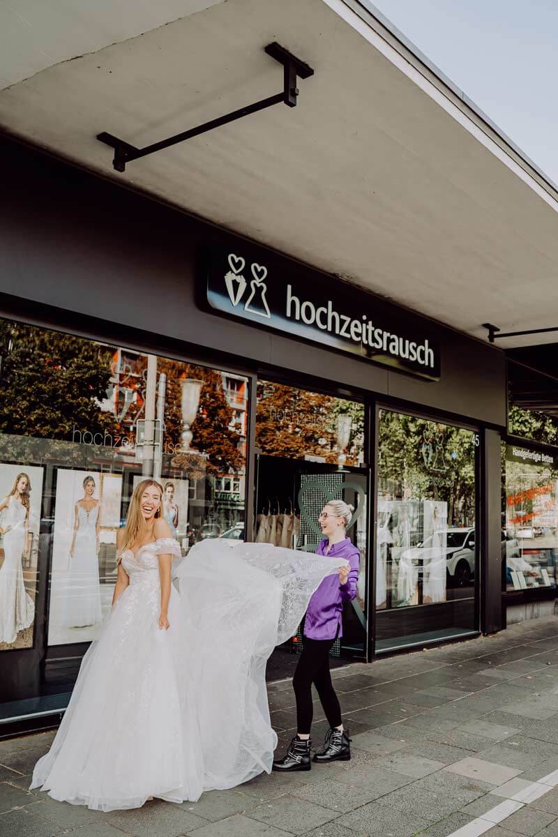 Eine lächelnde Braut in einem weißen Brautkleid steht vor dem hochzeitsrausch-Brautgeschäft, während eine andere Person die Schleppe anhebt. Im Schaufenster dahinter sind elegante Brautmoden und Hochzeitskleider ausgestellt.