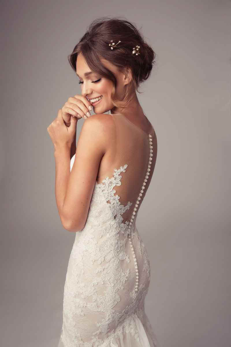 Eine lächelnde Braut in einem spitzenbesetzten Hochzeitskleid mit durchsichtigem Rücken und dekorativen Knöpfen steht im Profil, ihr Haar in einer Hochsteckfrisur mit kleinen floralen Akzenten, vor einem neutralen Hintergrund.