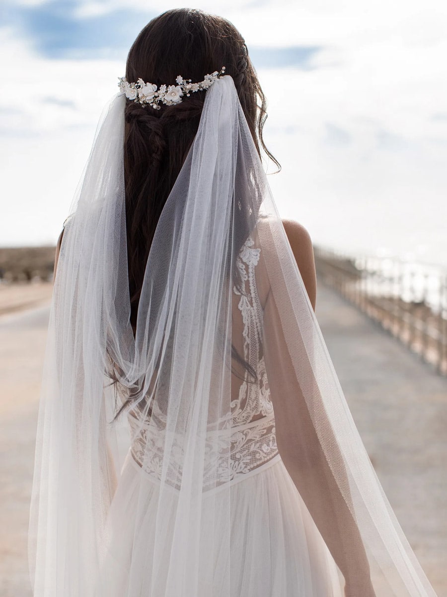 Eine Braut mit langem, dunklem Haar trägt das Charisse Brautkleid von Pronovias, ein zartes Kleid, das mit einem floralen Haarteil und einem Schleier kombiniert ist. Sie steht im Freien auf einem sonnenbeschienenen Weg am Meer - eine ideale Inspiration für moderne Brautmode.