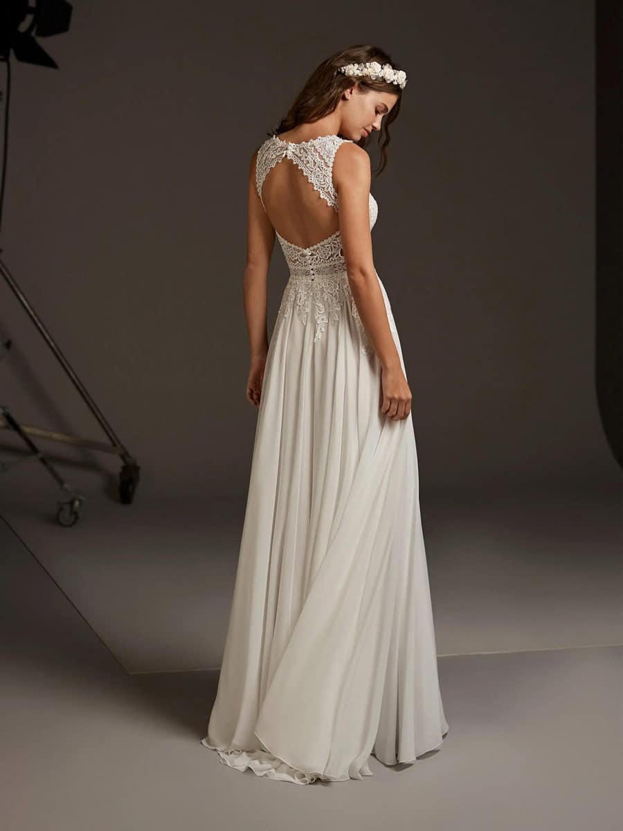 Eine Frau im Comet-Hochzeitskleid von Pronovias - ein langes, weißes Kleid mit offenem Rücken - steht mit einem geblümten Stirnband vor einer sanft beleuchteten Studiokulisse und schaut über ihre Schulter. Die perfekte Inspiration für die Braut.