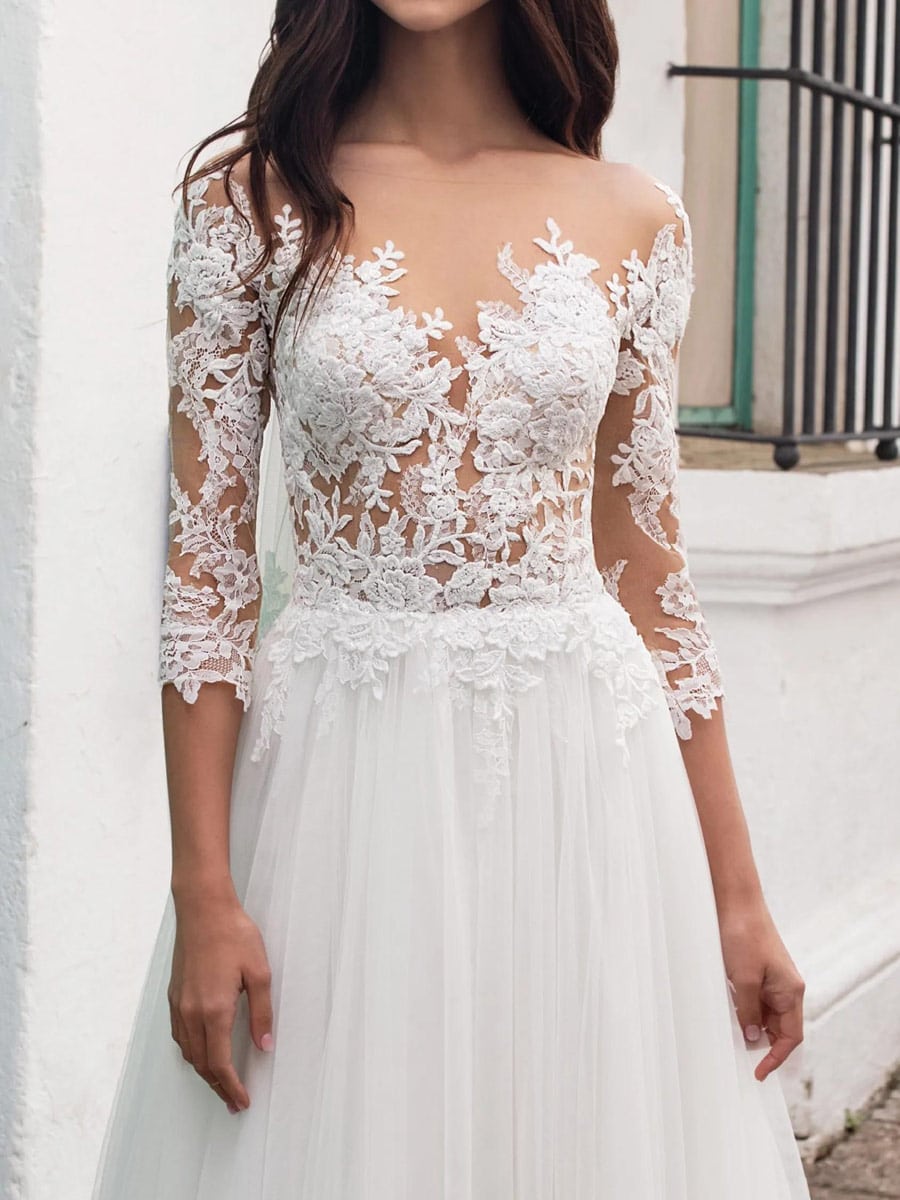 Eine Frau in einem Fornax-Brautkleid von Pronovias mit durchsichtigen langen Ärmeln und aufwändiger Blumenspitze am Mieder und an den Ärmeln, die im Freien vor einer weißen Wand steht.