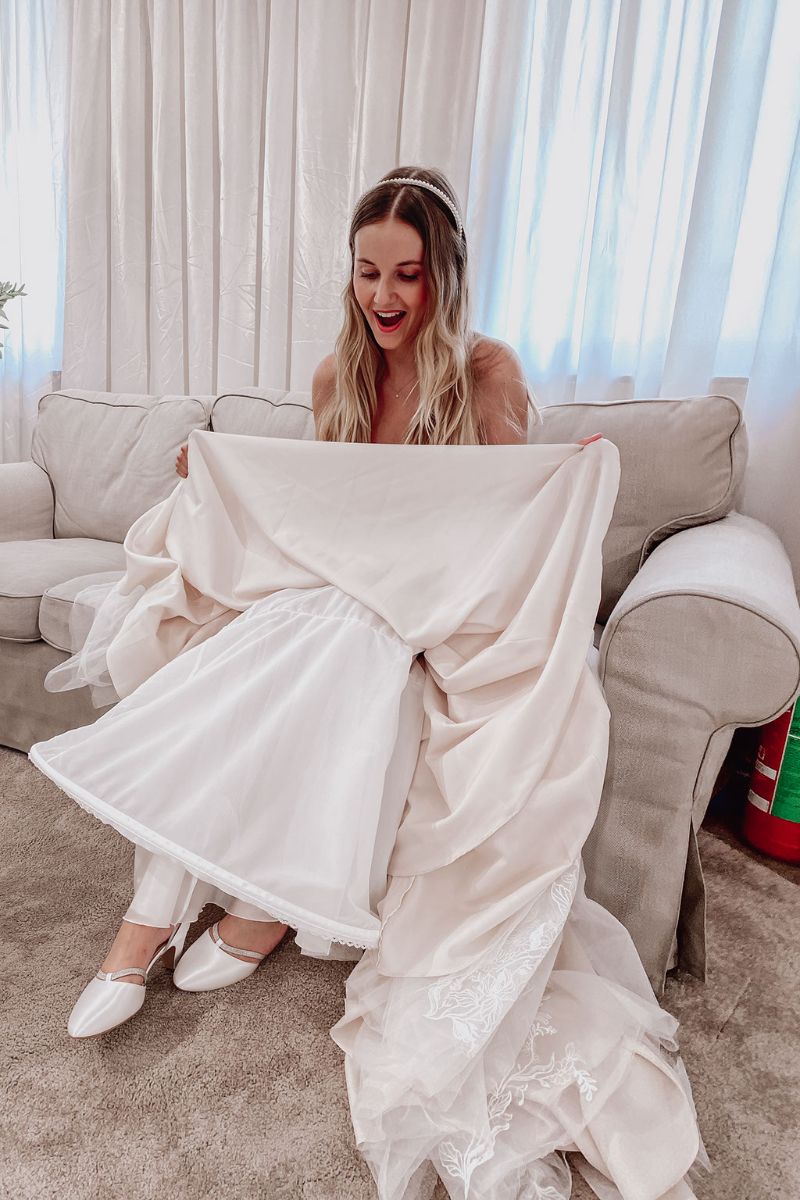 Eine lächelnde Braut sitzt auf einer hellen Couch und hebt ihr Hochzeitskleid an, so dass darunter eine weiße Hose und flache Schuhe zum Vorschein kommen. Der Raum ist sanft beleuchtet, mit weißen Vorhängen im Hintergrund.