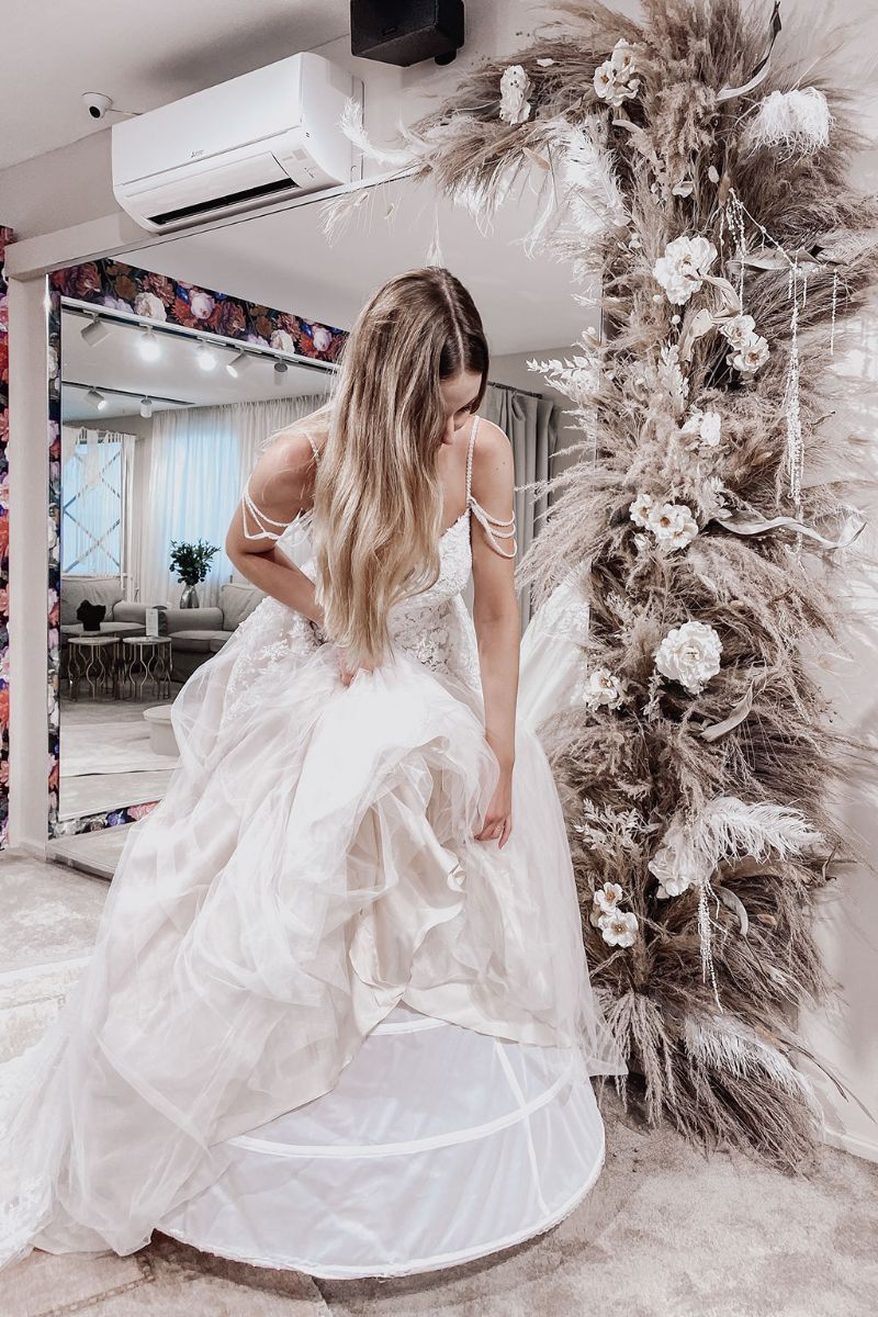 Eine Frau in einem weißen Brautkleid rückt die Lagen ihres Kleides in einem stilvollen Raum mit einem großen Spiegel, Blumenarrangements und Pampasgrasdekor zurecht. Natürliches Licht erfüllt den Raum und unterstreicht die Eleganz der modernen Brautmoden.