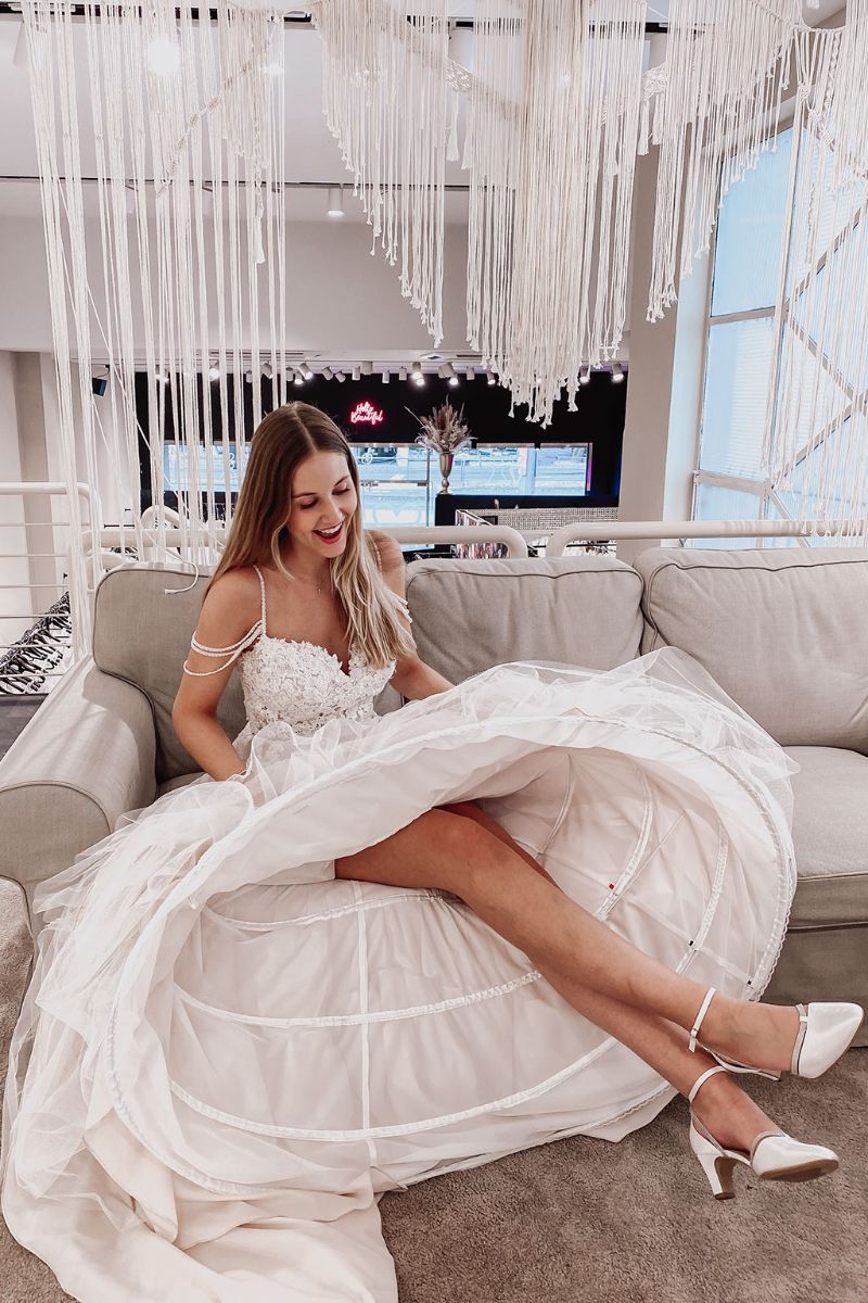 Eine lächelnde Frau in einem weißen Brautkleid mit Blumendetails sitzt auf einem Sofa und hebt ihren Rock leicht an, um die darunter liegenden Schichten zu enthüllen. Sie trägt weiße Absätze und ist von eleganten, hängenden weißen Kordeldekorationen umgeben.