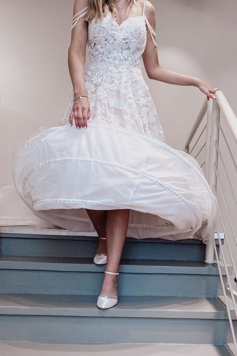 Eine Frau in weißem Brautkleid und weißen Schuhen geht eine Treppe hinunter und hebt dabei ihren Rock an, so dass darunter ein Unterrock zum Vorschein kommt. Das Bild ist über ihrem Mund abgeschnitten und konzentriert sich auf ihr unteres Gesicht und ihr elegantes Hochzeitskleid.