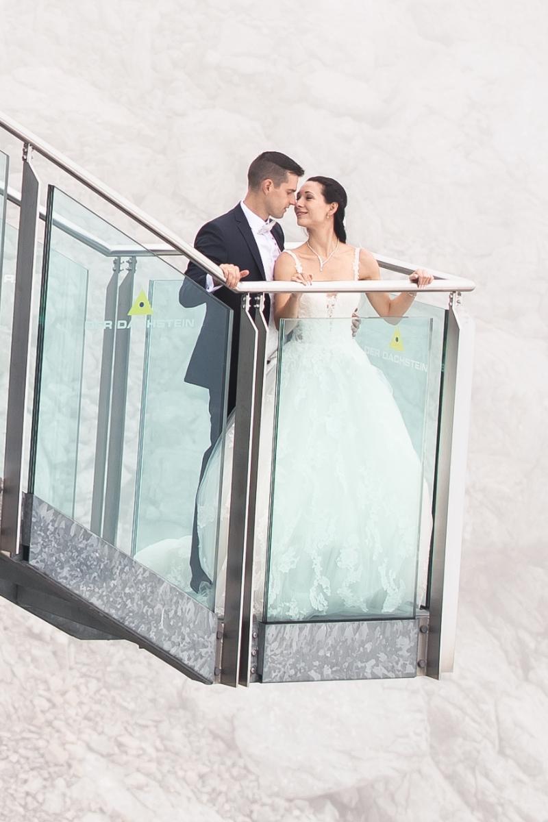 Eine Braut in einem atemberaubenden Brautkleid und ihr Bräutigam stehen zusammen auf einem Glasbalkon und schauen sich liebevoll an. Der neblige oder verschneite Hintergrund unterstreicht die verträumte Atmosphäre dieses magischen Moments.