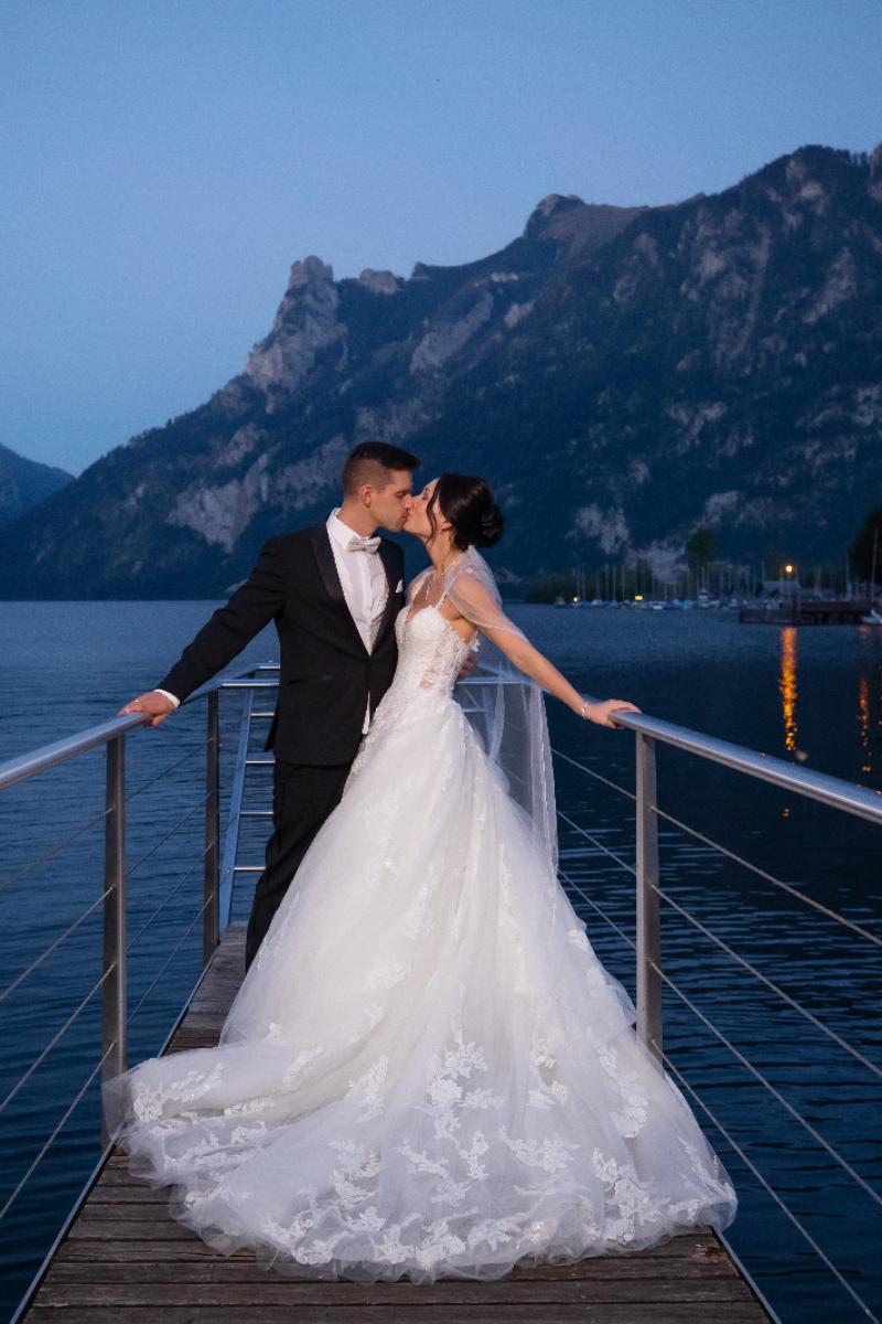 Eine Braut in einem atemberaubenden Hochzeitskleid und ihr Bräutigam küssen sich auf einem Steg über einem See in der Abenddämmerung, mit majestätischen Bergen im Hintergrund. Die Szene ist romantisch und heiter.
