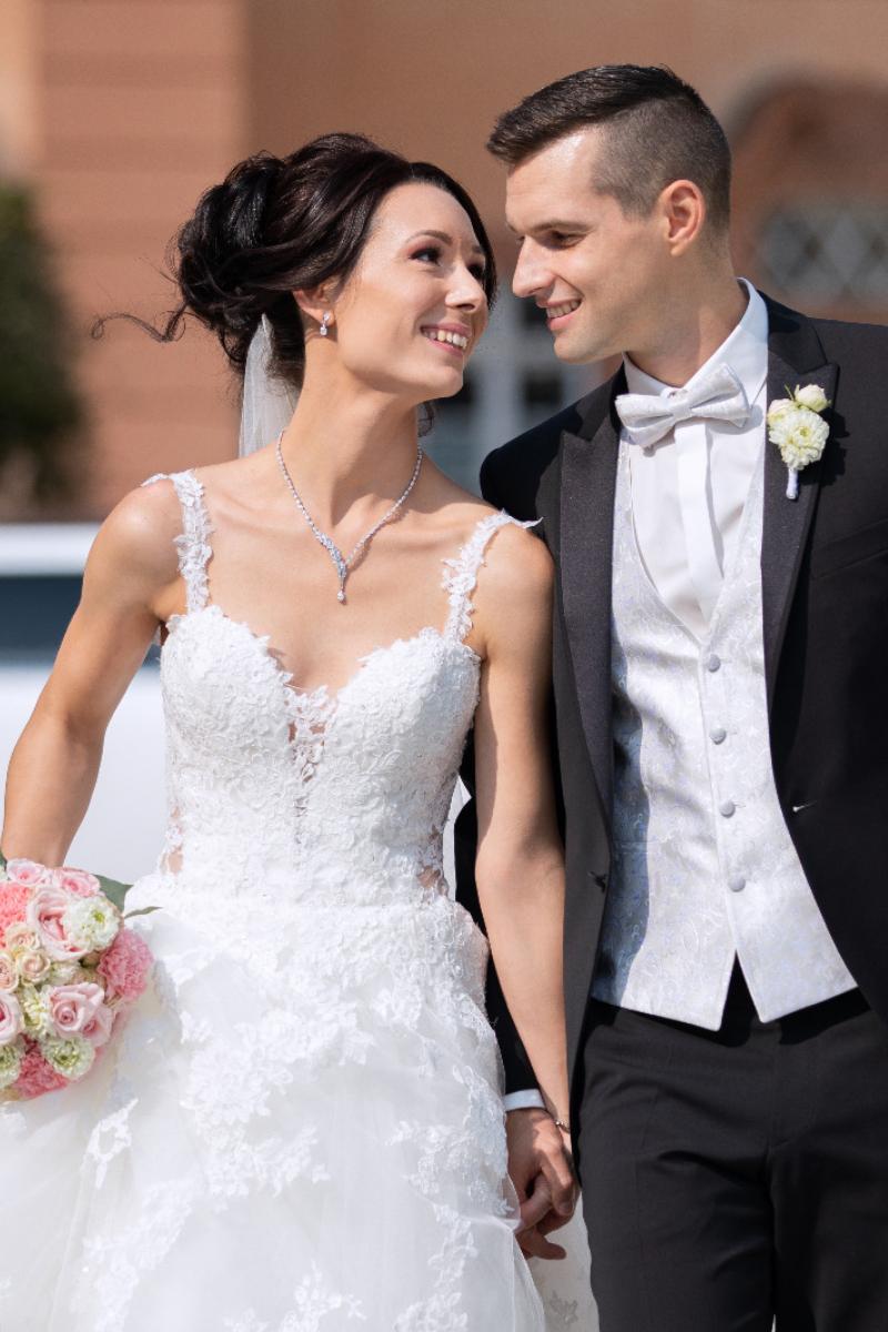 Eine Braut in einem weißen Spitzen-Hochzeitskleid mit einem Blumenstrauß steht neben einem Bräutigam im schwarzen Smoking mit weißer Weste und Fliege. Sie lächeln sich im Freien an und halten sich an den Händen, um ihren besonderen Tag in atemberaubenden Brautmoden zu feiern.