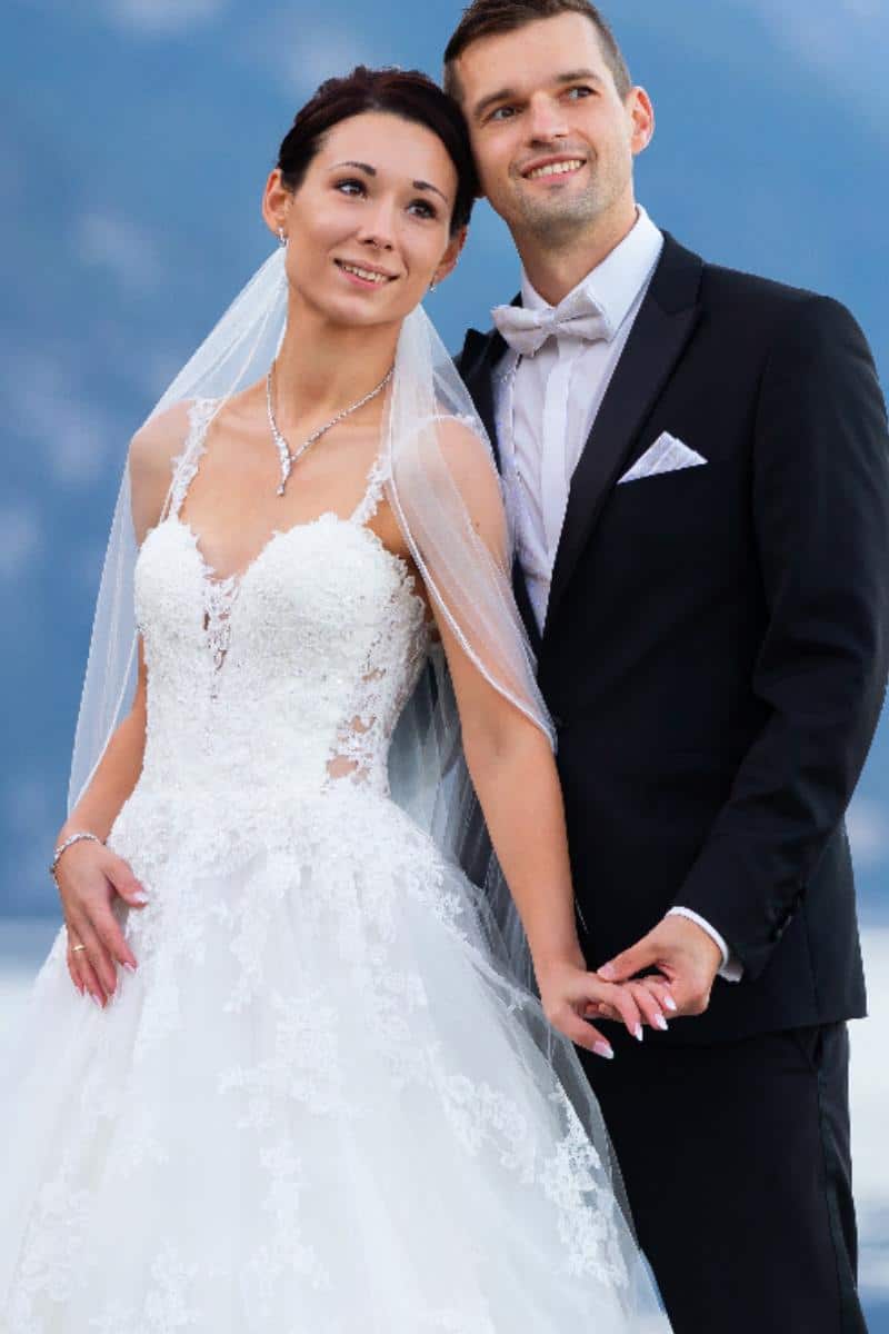 Eine Braut in einem weißen Spitzen-Brautkleid und Schleier steht neben einem Bräutigam in einem schwarzen Smoking und Fliege. Sie halten sich an den Händen und lächeln, wobei sie leicht nach oben blicken, mit einem unscharfen blauen Hintergrund im Hintergrund.