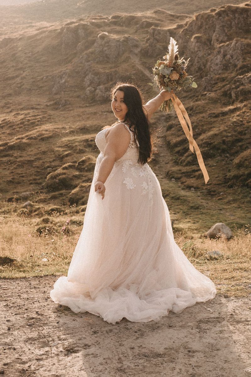 Eine lächelnde Braut in einem weißen Brautkleid hält einen Blumenstrauß in die Höhe, während sie im Freien auf einem sonnenbeschienenen, grasbewachsenen Hügel mit felsigem Gelände im Hintergrund steht.