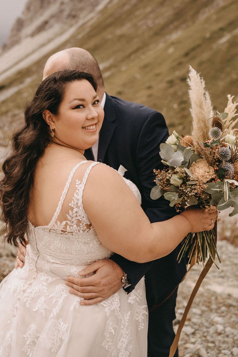Eine Braut im Brautkleid hält einen Strauß getrockneter Blumen und Grünzeug und lächelt, während ihr Bräutigam im dunklen Anzug sie im Freien umarmt, mit den Bergen im Hintergrund.