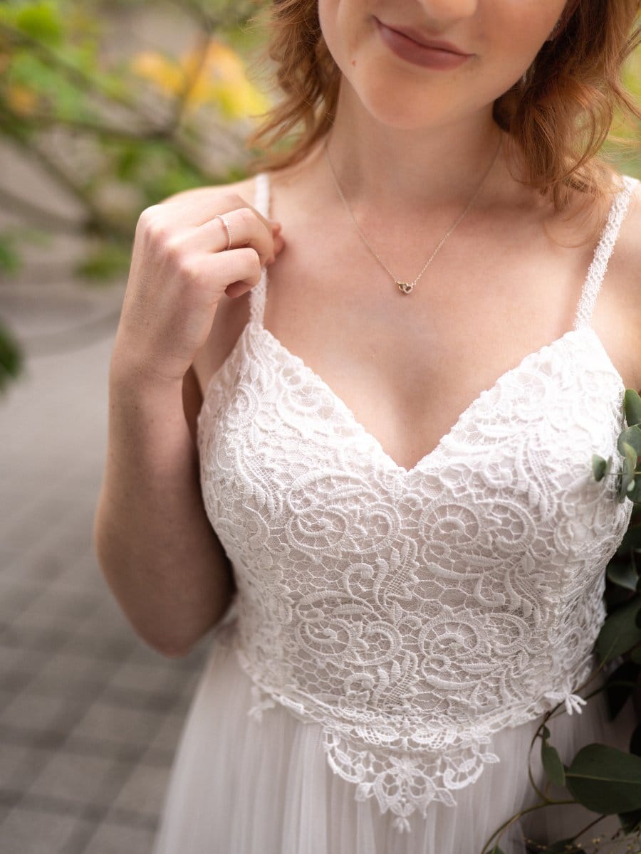 Eine Frau in einem weißen Spitzenbrautkleid hält einen Träger und einen Strauß Grünzeug. Sie trägt eine zarte Halskette und steht mit einem sanften Lächeln im Freien. Das Bild hebt ihren Oberkörper und die filigranen Details des Hochzeitskleides hervor.
