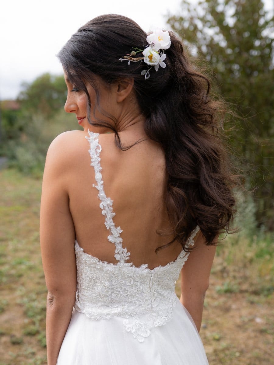 Eine Frau in einem weißen Spitzenbrautkleid steht im Freien mit dem Rücken zur Kamera. Ihr langes dunkles Haar ist zu lockeren Locken gestylt und mit weißen Blumen geschmückt, die sie an der Seite festgesteckt hat, vor einem verschwommenen Grün im Hintergrund.