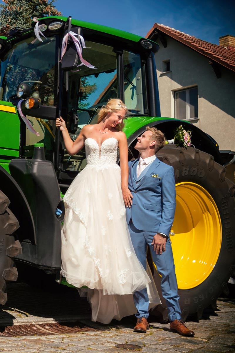 Eine Braut in einem atemberaubenden Brautkleid und ein Bräutigam in einem blauen Anzug stehen lächelnd neben einem großen grünen Traktor, der mit Bändern geschmückt ist, an einem sonnigen Tag in der Nähe eines Gebäudes.