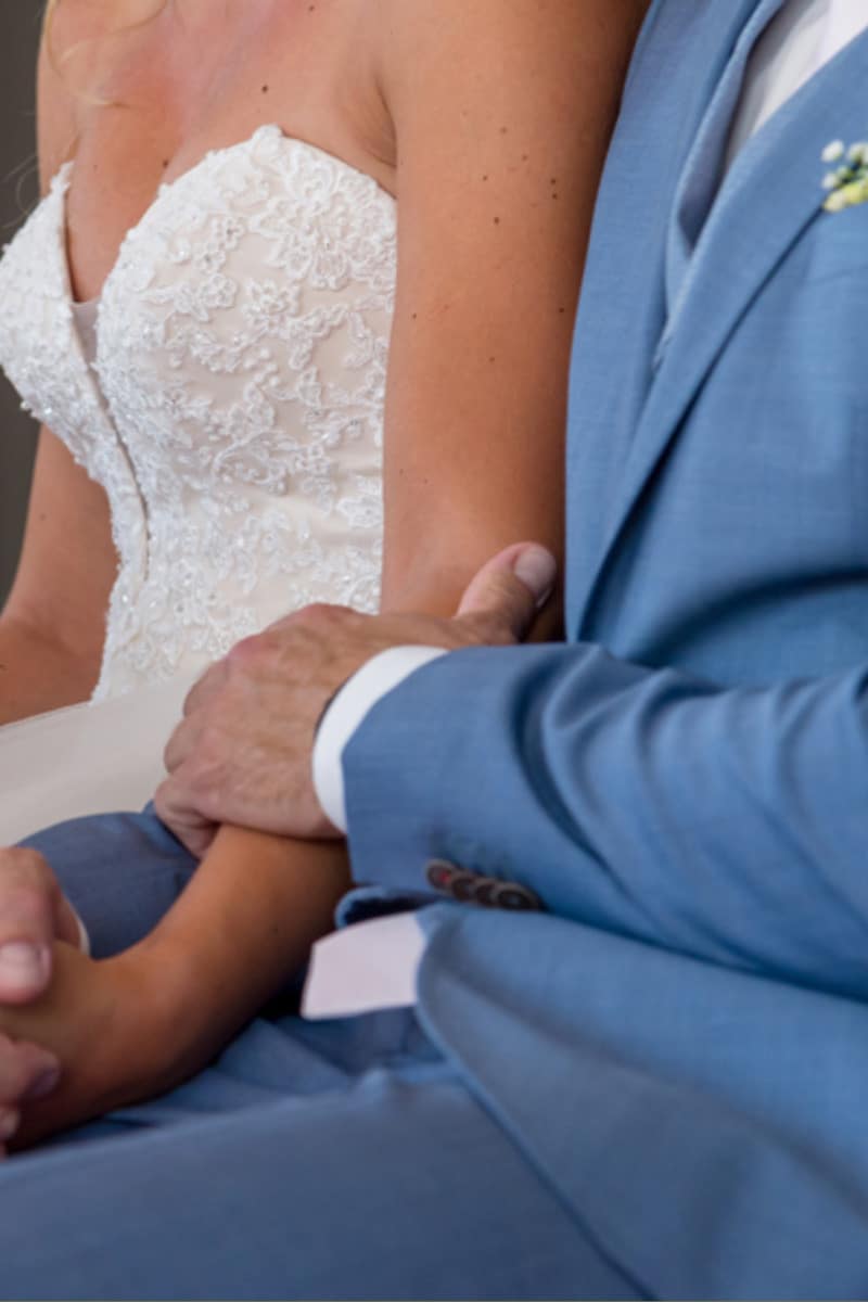 Eine Braut in einem weißen Spitzen-Hochzeitskleid sitzt neben einem Bräutigam in einem hellblauen Anzug und hält die Arme sanft ineinander verschränkt.