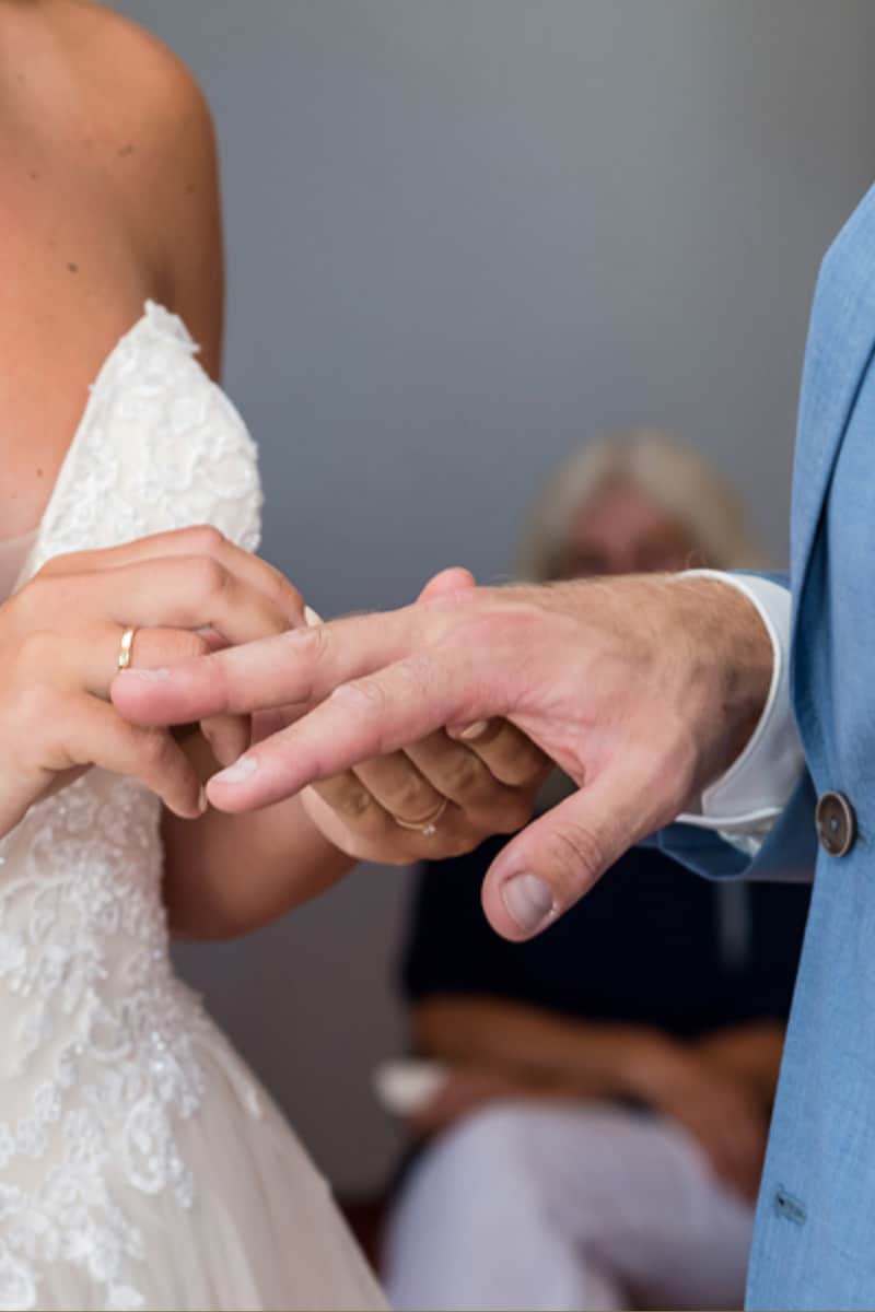 Eine Braut in einem weißen Hochzeitskleid steckt dem Bräutigam während einer Hochzeitszeremonie einen Ring an den Finger; beide sind von den Schultern abwärts dargestellt.