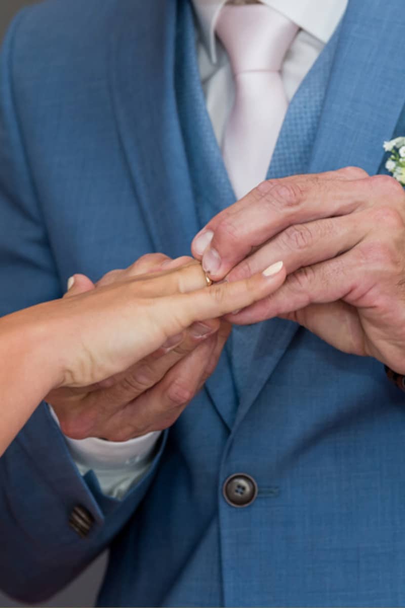 Eine Person in einem blauen Anzug steckt einer anderen einen Ring an den Finger, während die Braut elegant in ihrem Brautkleid steht, was auf eine Hochzeit oder Verlobungsfeier hindeutet.