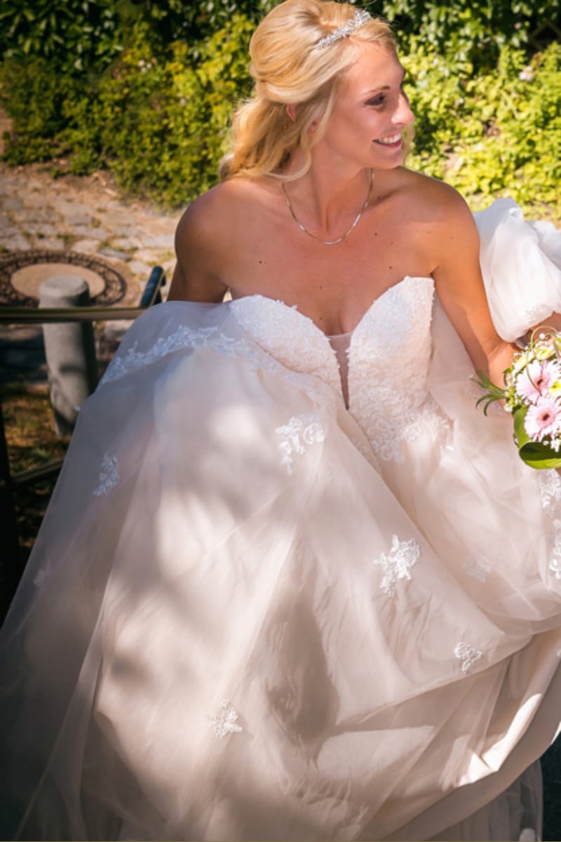 Eine lächelnde Braut in einem weißen, trägerlosen Brautkleid mit Spitzendetails hält ihr Kleid und einen Strauß rosafarbener Blumen in der Hand, während sie im Freien mit Grünpflanzen im Hintergrund steht.