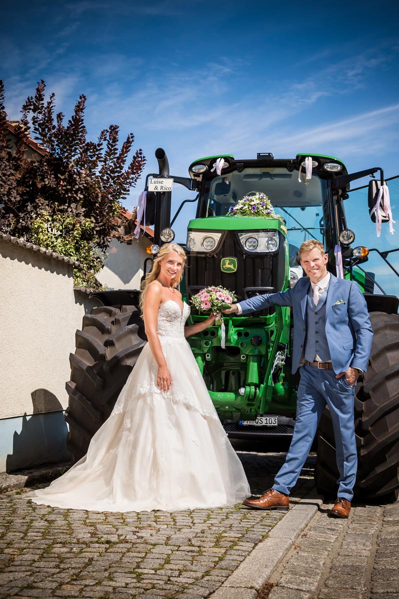Eine Braut in einem umwerfenden Brautkleid und ein Bräutigam in einem blauen Anzug posieren lächelnd vor einem grünen, mit Blumen und Bändern geschmückten Traktor an einem sonnigen Tag.