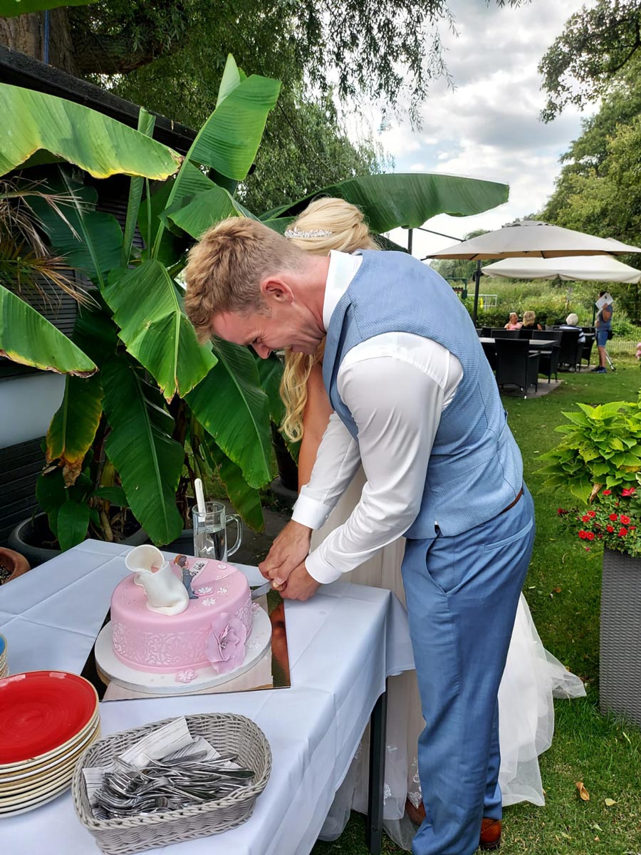 Eine Braut in einem umwerfenden Brautkleid und ein Bräutigam in einem hellblauen Anzug schneiden im Freien eine rosa Hochzeitstorte an, umgeben von üppigem Grün und großen Blättern. Ihre eleganten Brautmoden bildeten den Rahmen für diese schöne Feier.