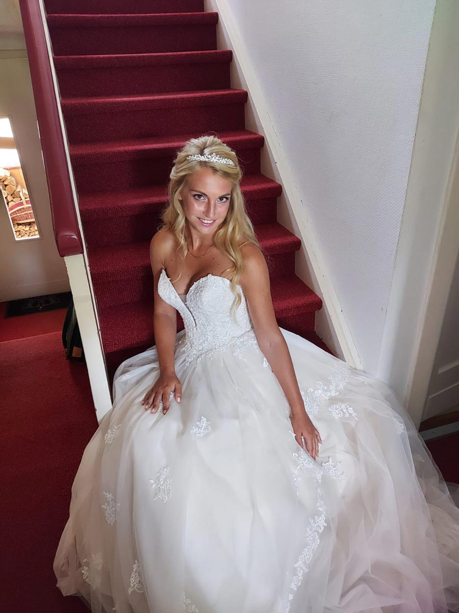 Eine Braut in einem weißen Spitzenbrautkleid und Diadem sitzt auf einer Treppe mit rotem Teppich und lächelt in die Kamera.