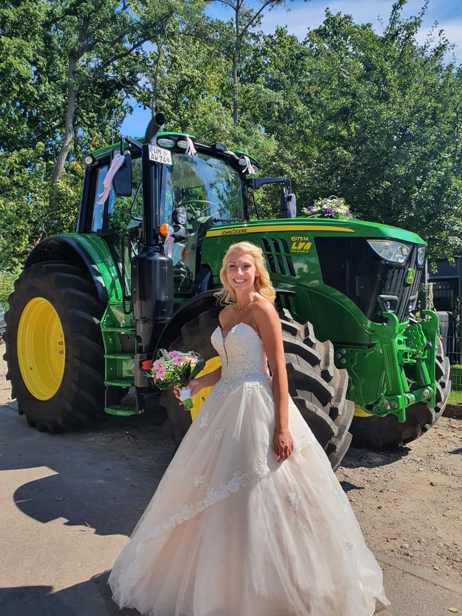 Eine lächelnde Braut in einem eleganten Brautkleid steht draußen mit einem Blumenstrauß vor einem großen grünen Traktor, der mit Bändern geschmückt ist. Sonnenlicht und Bäume bilden die Kulisse für diesen besonderen Brautmoden-Moment.