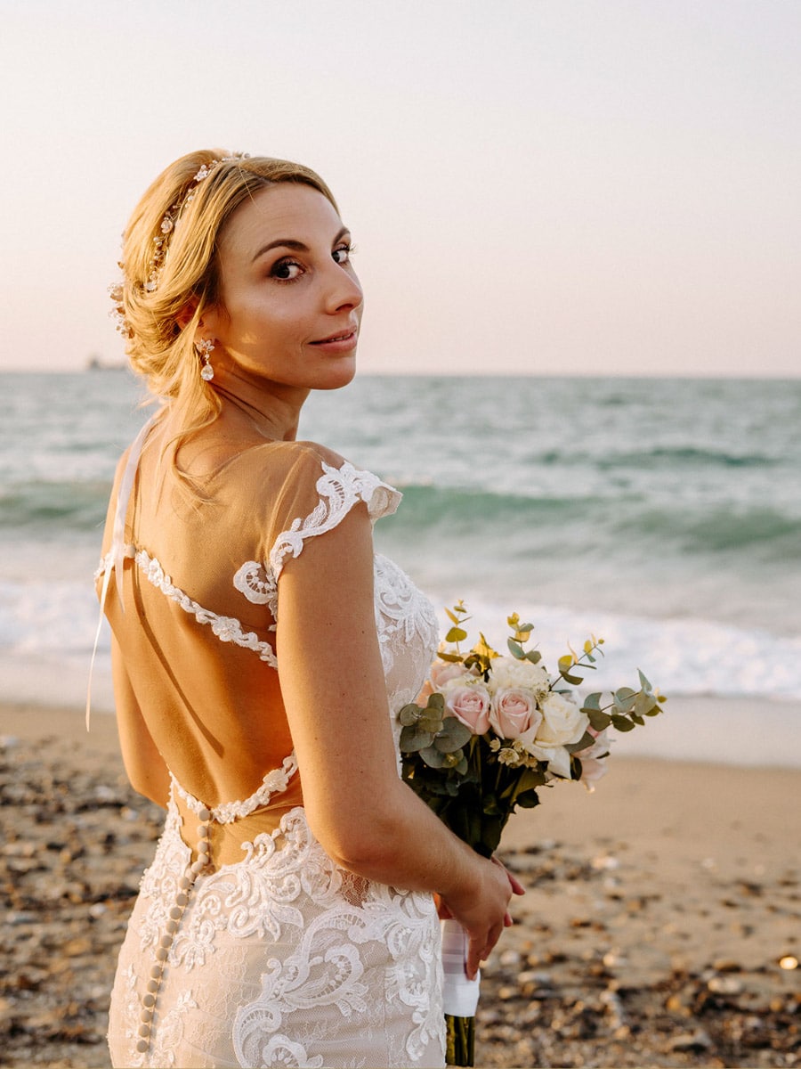 Eine Braut in einem zarten Hochzeitskleid steht am Strand und hält einen Strauß aus Rosen und Grünzeug in der Hand. Sie blickt über ihre Schulter in Richtung Kamera, eingerahmt vom Meer und dem Sonnenuntergang im Hintergrund.