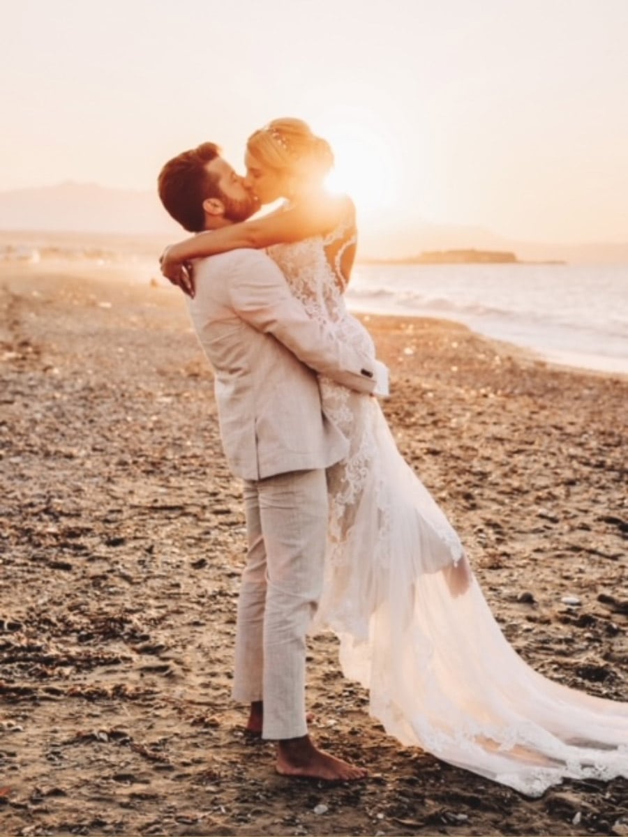 Ein frisch verheiratetes Paar umarmt und küsst sich an einem Sandstrand bei Sonnenuntergang. Das elegante Brautkleid der Braut leuchtet im goldenen Licht, während der helle Anzug des Bräutigams die romantische Szene vervollständigt, während die Sonne hinter ihnen untergeht.