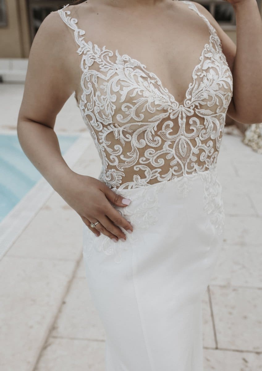 Eine Frau steht am Pool und trägt das Bridentity Admiration Hochzeitskleid: ein ärmelloses, weißes Brautkleid mit transparenter, bestickter Spitzenpasse und figurbetontem Rock - zeitlos elegant und modern.