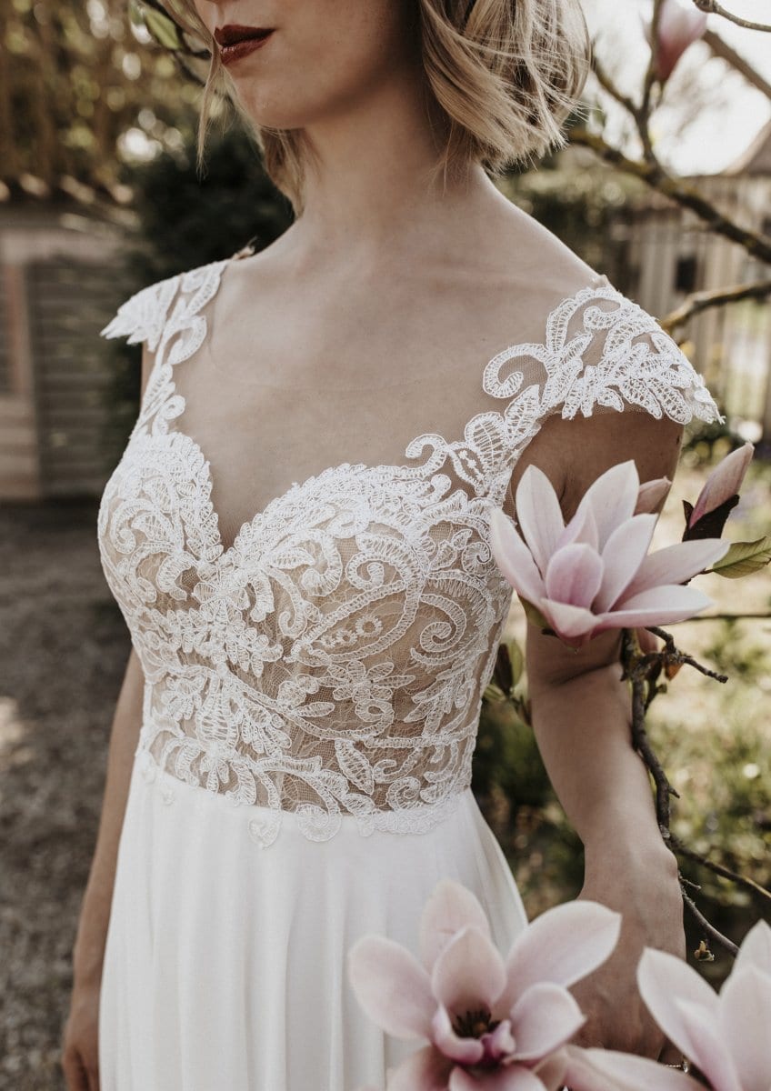 Eine Frau im Bridentity Affection Brautkleid, einem ärmellosen weißen Kleid mit aufwendigen Blumendetails, steht im Freien und hält einen blühenden Magnolienzweig, der eine sanfte und romantische Szene schafft.