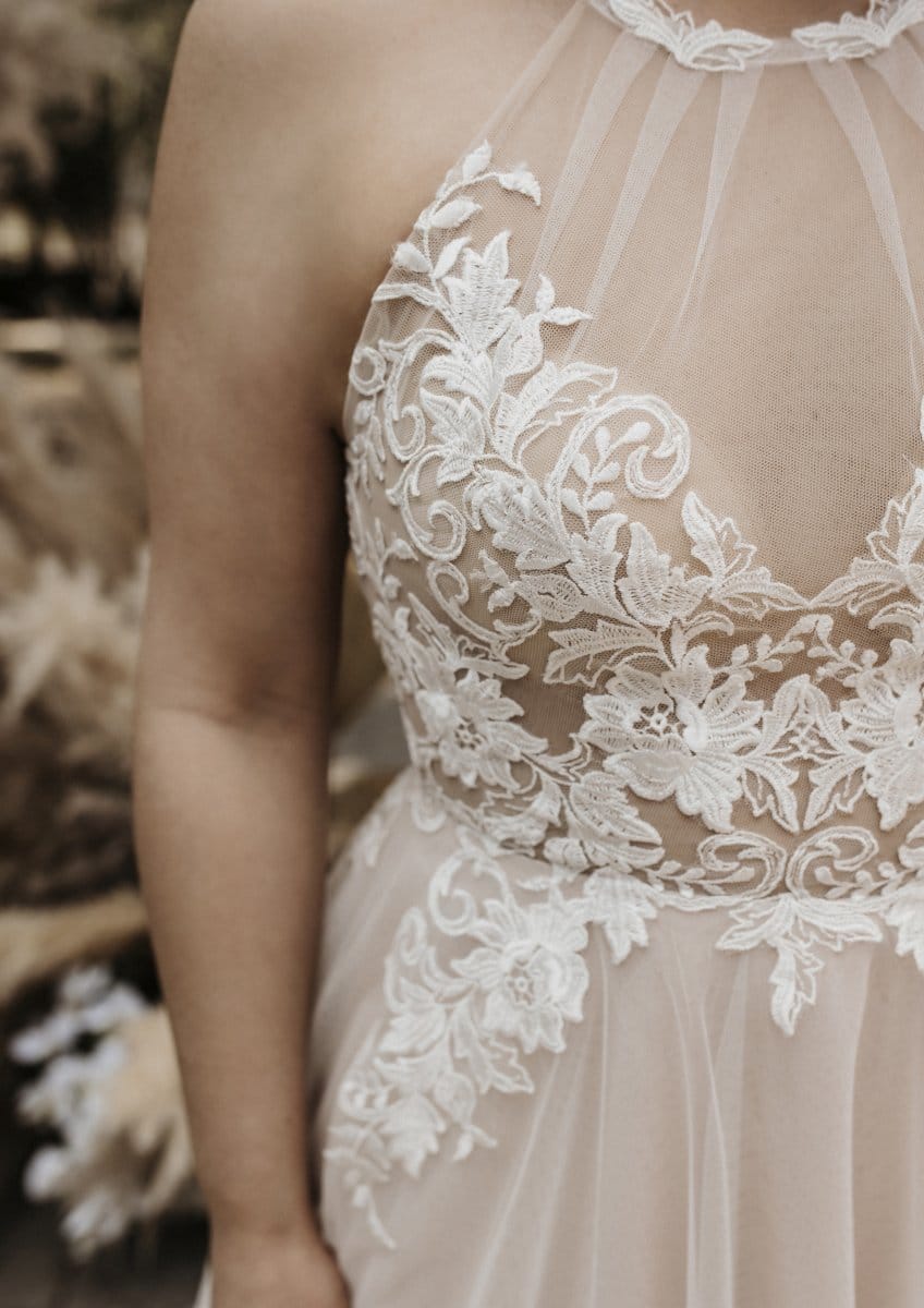 Nahaufnahme einer Person, die das Bridentity Bliss Brautkleid trägt, ein ärmelloses, durchsichtiges Hochzeitskleid mit aufwändiger weißer, floraler Spitzenstickerei auf dem Mieder.