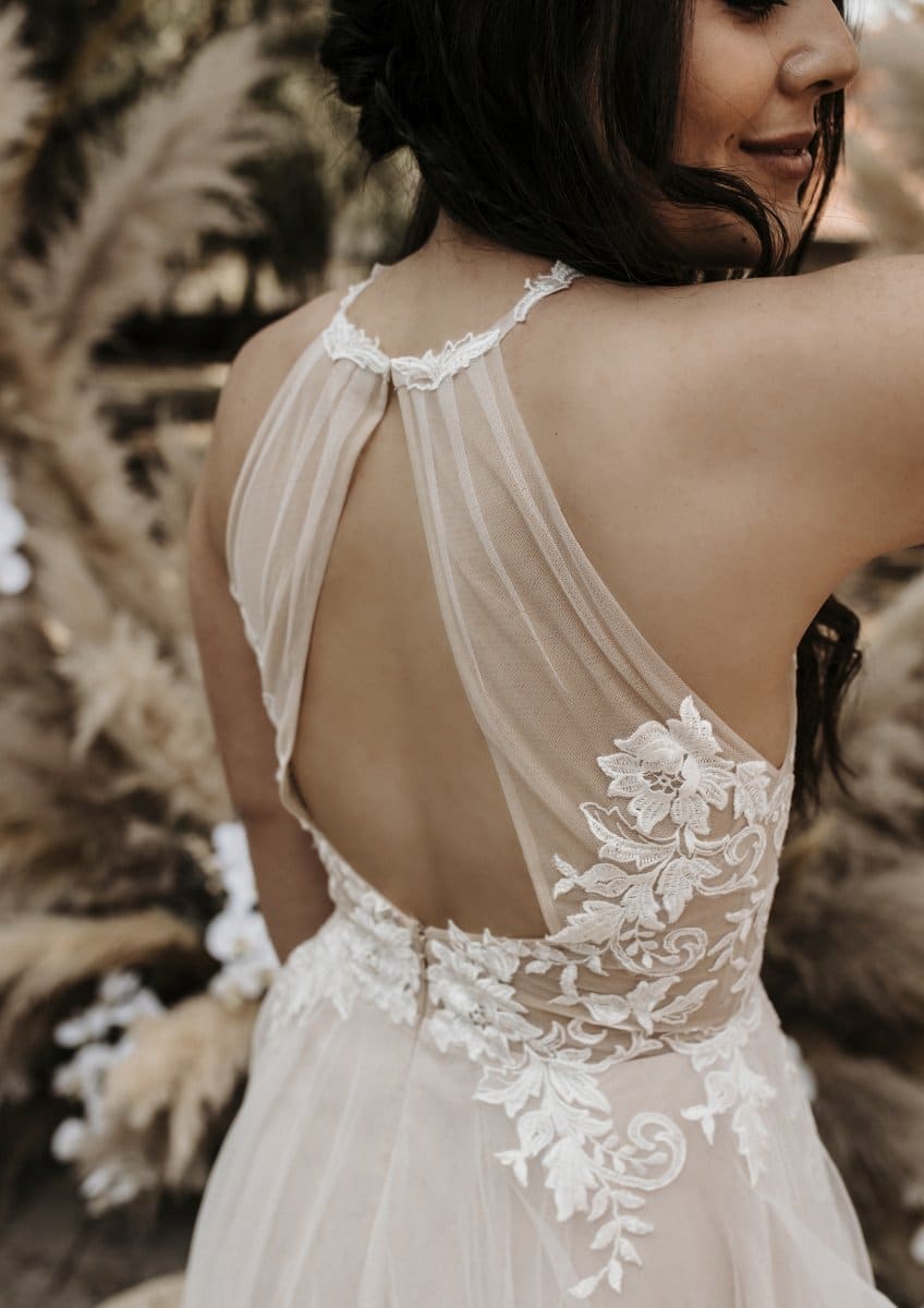 Eine Frau in einem ärmellosen, rückenfreien Bridentity Bliss Brautkleid mit floraler Spitzenstickerei steht im Freien und lächelt sanft. Ihr dunkles Haar fällt in lockeren Wellen, und Pampasgras akzentuiert den unscharfen, von einer Braut inspirierten Hintergrund.