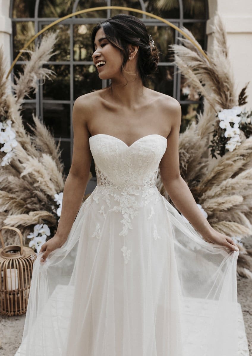 Eine lächelnde Braut trägt das trägerlose, spitzenverzierte Bridentity Charm Hochzeitskleid im Freien und hält ihr Kleid. Der Hintergrund ist mit Pampasgras und weißen Blumen geschmückt.