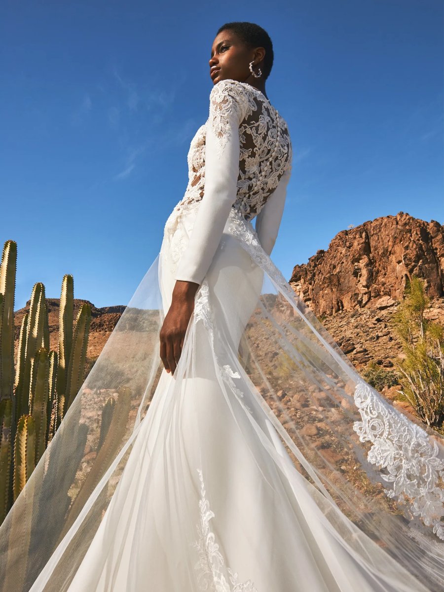 Eine Frau in einem taillierten, langärmeligen Pronovias Dazu-Hochzeitskleid steht unter freiem Himmel zwischen Kakteen und Felsen, die Schleppe ihres Kleides fließt hinter ihr her und der Himmel ist blau.