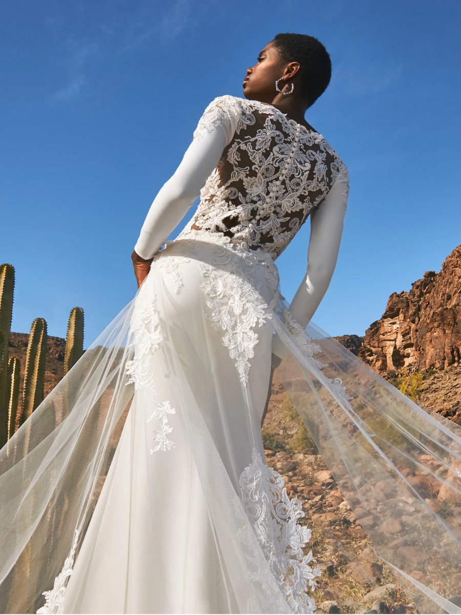 Eine Frau im Pronovias Dazu Hochzeitskleid mit transparenten Blütendetails steht im Freien zwischen Kakteen und felsigen Hügeln und blickt in den strahlend blauen Himmel.