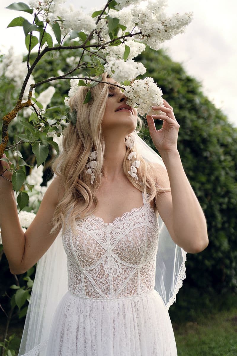 Eine Braut im Dama Couture Bjork Brautkleid steht im Freien, schnuppert an weißen Blüten und ist umgeben von sattem Grün sowie wolkigem Himmel - eine romantische Inspiration für Brautmode.