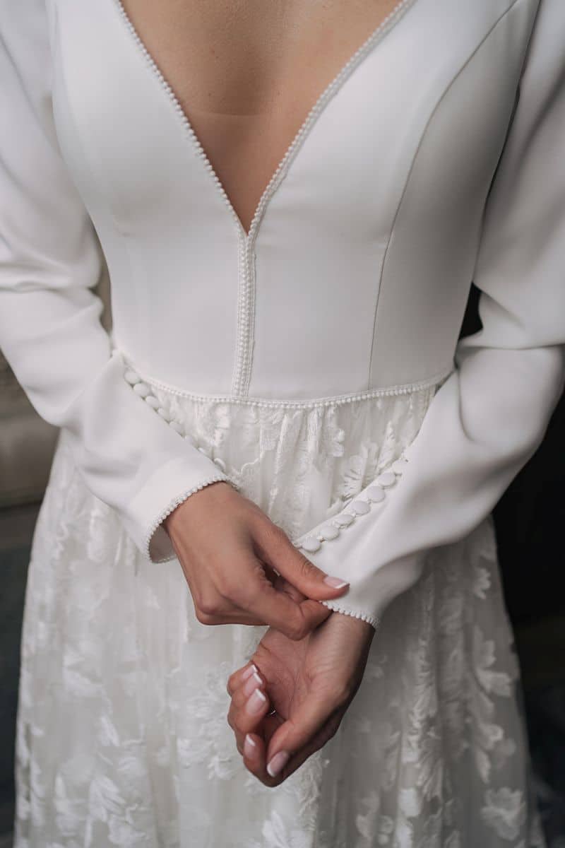 Eine Frau in dem Dama Couture Latifa Hochzeitskleid mit langen Ärmeln und tiefem V-Ausschnitt rückt die geknöpfte Manschette am Ärmel zurecht. Das taillierte Mieder und der Rock aus floraler Spitze unterstreichen den raffinierten Stil dieses eleganten Brautkleides.
