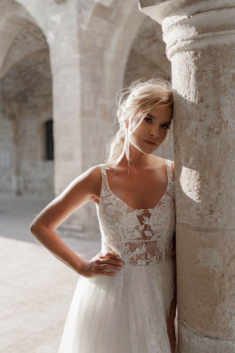 Eine Frau in einem ärmellosen Dama Couture Rosa Brautkleid lehnt an einer Steinsäule in einem sonnendurchfluteten, gewölbten Innenhof, wobei das sanfte Sonnenlicht ihr Gesicht und ihr Haar beleuchtet.