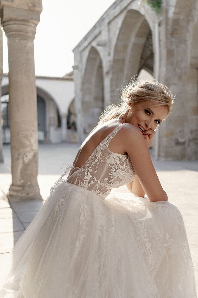 Eine lächelnde Frau im Dama Couture Rosa Brautkleid sitzt im Freien unter steinernen Bögen, das Sonnenlicht beleuchtet sanft ihr Gesicht und ihr Kleid - und unterstreicht die zeitlose Eleganz dieses Spitzenbrautkleides.